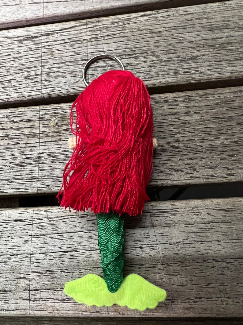 May include: A red haired mermaid keychain with a green tail and light green fins. The keychain has a silver ring.