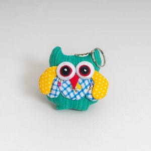 Op de afbeelding: Een groene stoffen uil sleutelhanger met gele stippen op de vleugels en rode ogen. De uil heeft een blauw-wit geruit patroon op zijn borst.