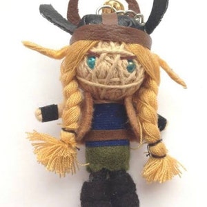 May include: A keychain doll featuring a character with blonde yarn hair in two braids, wearing a brown leather-like helmet with horns. The doll has blue bead eyes, a blue and green fabric torso, black cuffs, and black fabric boots with a metal ring.