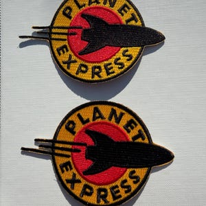May include: Two circular embroidered patches featuring a red center circle surrounded by a yellow border. A black rocket silhouette extends horizontally across the center. The text PLANET EXPRESS is stitched in black along the yellow border.
