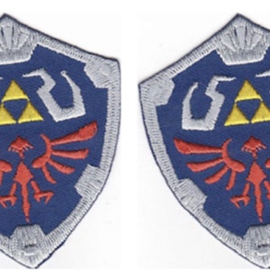May include: Two identical shield-shaped embroidered patches featuring a blue background, a central yellow triforce symbol, a red bird-like emblem below, and silver-grey border detailing with a small crown-like motif at the top of each shield.