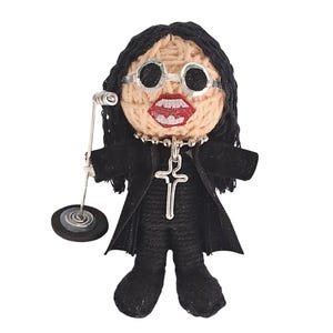Op de afbeelding: Handgemaakte pop van een rockster, met zwart haar, een ronde zilveren bril en een zwarte outfit. De pop heeft een zilveren kruis ketting en houdt een zilveren microfoon vast. Het gezicht van de pop is gedetailleerd met rode lippen.
