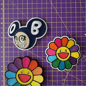 May include: Three embroidered patches on a purple cutting mat with a grid. One patch is a dark blue character head with white ears and the letter B. Two patches are multi-coloured flowers with yellow centres and rainbow petals in a circular pattern.