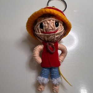 Op de afbeelding: Handgemaakte sleutelhanger met een garend popje dat lijkt op een personage uit de anime-serie One Piece. De pop heeft een strohoed met rode en gele accenten, een rood vest, een blauwe spijkerbroek en armen en benen van garen. De sleutelhanger is bevestigd aan een zilveren metalen ring.