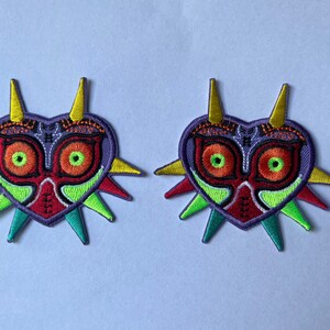 以下が含まれることがあります： ハート型のマスクをデザインした、同一の刺繍ワッペン2枚。マスクは主に紫色で、大きなオレンジ色と緑色の目、上部に黄色のスパイク、側面には赤、緑、青緑色のスパイクが施されています。
