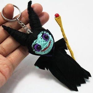 Puede incluir: Una muñeca de lana negra con una cara azul y verde, que lleva una capa negra y sostiene una varita dorada. La muñeca está unida a un llavero plateado.