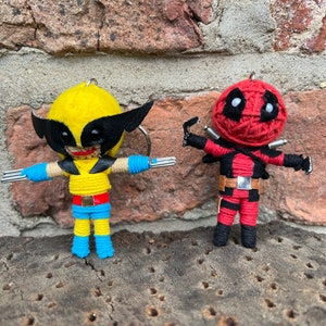 Puede incluir: Dos figuras de arte de hilo de personajes populares de cómics, Wolverine y Deadpool. Wolverine es amarillo con garras negras y un traje azul y amarillo. Deadpool es rojo con ojos negros y un cinturón negro.