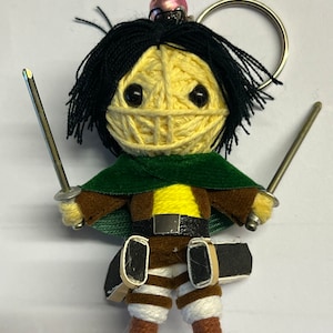 Puede incluir: Un llavero de muñeca de hilo hecho a mano con un personaje de pelo negro, capa verde y ropa marrón y blanca. La muñeca sostiene dos espadas plateadas y tiene una cara amarilla. Una campana rosa y un llavero están adjuntos.