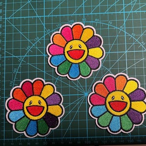Könnte beinhalten: Drei gestickte Blumenaufnäher, jeder mit einem gelben Gesicht in der Mitte, einem breiten roten Lächeln und schwarzen Augen. Die Blütenblätter um die Mitte sind in einem Regenbogenmuster aus Rot, Orange, Gelb, Grün, Blau und Violett angeordnet.