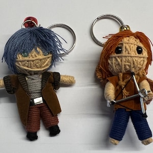 Puede incluir: Dos llaveros de muñecas hechos a mano. Una muñeca tiene pelo azul, una chaqueta marrón y un cinturón plateado. La otra muñeca tiene pelo rojo, un chaleco marrón y pantalones azules. Ambas muñecas tienen caras de hilo y sostienen accesorios.