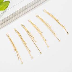 Könnte beinhalten: Sechs goldfarbene Schmuckanhänger mit einem mehrschichtigen Kettendesign und kleinen, hängenden Perlen. Jeder Anhänger hat oben eine kleine Schlaufe zum Befestigen an anderen Schmuckkomponenten. Die Anhänger sind diagonal auf einer weißen Oberfläche angeordnet.