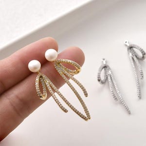 May include: Two gold-toned earrings featuring a white spherical pearl stud attached to a dangling, elongated, ribbon-like charm encrusted with small clear crystals. Two additional silver-toned ribbon charms with clear crystals are positioned nearby.