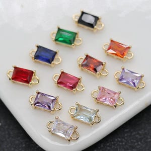 May include: Various rectangular gemstone charms in colours such as red, green, blue, and pink. Each charm has a gold-coloured frame and small loops for attachment. The gemstones are arranged on a white surface.