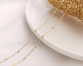 May include: Close-up of a spool of gold-colored chain with small, round beads and rectangular links. The chain is displayed on a white plate, suggesting a jewelry-making or craft supply.