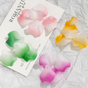 Peut inclure: Un assortiment de perles translucides en forme de pétales, dans des tons de rose, blanc, jaune et vert. Chaque perle possède un petit trou pour l'enfilage. Les perles sont disposées sur une surface blanche avec une carte imprimée.