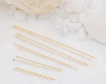 May include: Gold-colored head pins of varying lengths, arranged on a white surface. The pins have a small, flat head and a long, slender shaft. The background includes white pebbles and a seashell, suggesting a craft or jewelry-making theme.