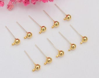 May include: A collection of gold-colored earring posts with a small ball at the end of each post. The posts are arranged on a white surface, with pink flowers in the background.