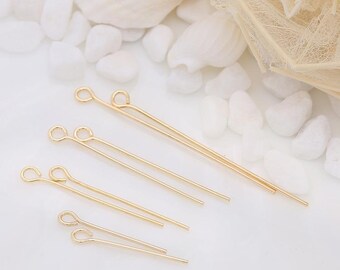 Può includere: Diverse spilli a occhiello color oro di varie lunghezze sono disposti su una superficie bianca. Gli spilli hanno un anello a un'estremità e una punta all'altra. Lo sfondo include ciottoli bianchi e una conchiglia.