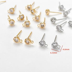 May include: A collection of gold and silver-colored stud earrings with clear, round gemstones. The earrings have a textured, cup-like setting and a small loop. Measurements are 6.5mm long and 4.6mm wide.