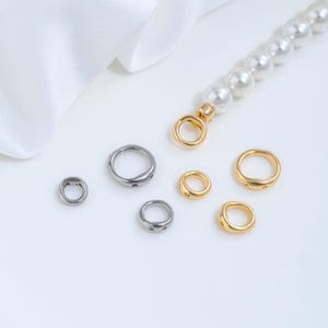 Può includere: Un assortimento di componenti per gioielli, tra cui anelli aperti color argento e oro. È visibile anche una collana di perle con chiusura dorata. I componenti sono disposti su una superficie bianca.