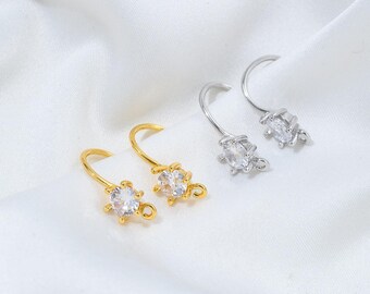 May include: Two pairs of earrings, one gold-toned and one silver-toned, are displayed. Each earring features a curved hook design with a clear, oval-shaped gemstone at the base. The gemstones are surrounded by a decorative setting.