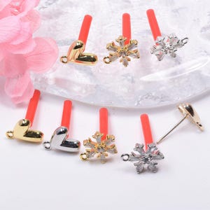 May include: A collection of decorative ear studs featuring heart and snowflake designs. The studs have red posts and are made of gold and silver-colored metal. The heart studs are in gold and silver, while the snowflake studs have crystal accents.