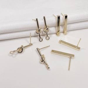 May include: Gold-toned metal earrings in various designs. The collection includes bar-shaped studs and earrings with a knotted design. The earrings are displayed on a white surface, showcasing their shiny finish and intricate details.