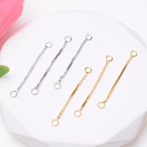 May include: An assortment of jewelry chain extenders in silver and gold tones. The extenders have a small ring at each end and are designed with different chain styles. The background is white with a hint of pink.