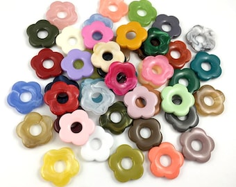 May include: A collection of flower-shaped beads in various colors, including pink, green, blue, and brown. The beads have a hole in the center and are made of a glossy material. The beads are scattered on a white surface.