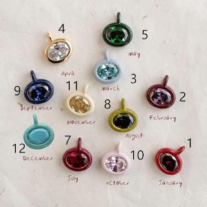 May include: Twelve oval-shaped pendants with faceted stones in various colours, each labelled with a number and a month. The collection includes clear, green, light blue, purple, dark blue, yellow, olive, turquoise, red, and pink stones in metal settings.
