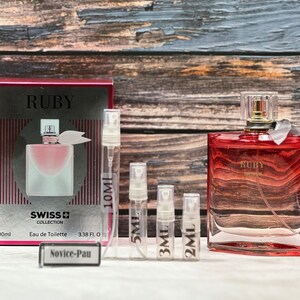 Puede incluir: Un frasco de perfume Ruby de vidrio rojo y transparente con una tapa dorada. El frasco está sobre una superficie de madera con un frasco de perfume más pequeño y una caja de perfume Ruby. La caja es roja y blanca y tiene el texto "Ruby Swiss Collection Eau de Toilette 100ml 3.38 fl oz". Los frascos más pequeños están etiquetados con el texto "10ml", "5ml", "3ml", y "2ml".