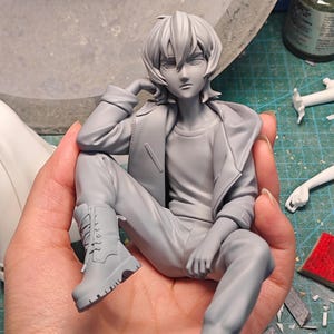 May include: A grey figurine of a person in a seated pose, wearing a jacket, trousers, and boots. The figure has short hair and is resting their arm on their knee. The boots have detailed laces and treads. The figurine is held in a hand.