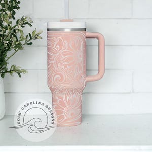 Peut inclure: Un gobelet isotherme en acier inoxydable rose avec un motif floral blanc et une poign&eacute;e noire. Le gobelet a un couvercle blanc et une paille. Le gobelet est pos&eacute; sur une surface blanche. Le texte "Goin' Carolina Designs" est visible dans le coin inf&eacute;rieur gauche de l'image.