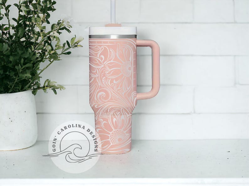 Peut inclure: Un gobelet isotherme en acier inoxydable rose avec un motif floral blanc et une poign&eacute;e noire. Le gobelet a un couvercle blanc et une paille. Le gobelet est pos&eacute; sur une surface blanche. Le texte "Goin' Carolina Designs" est visible dans le coin inf&eacute;rieur gauche de l'image.