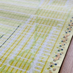 May include: Close-up of textured paper with a yellow and green grid pattern. The paper has a handmade appearance with visible fibres. Gold and navy blue accents are along the edge, adding a decorative touch. The paper is on a wooden surface.