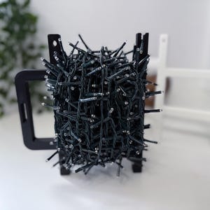 Könnte beinhalten: Ein dicht gewickeltes Bündel grüner Lichterketten mit kleinen weißen Glühbirnen wird von einem schwarzen Metallrahmen gehalten. Die Lichter sind dicht gepackt und erzeugen ein strukturiertes Aussehen. Der Rahmen hat ein griffartiges Design.