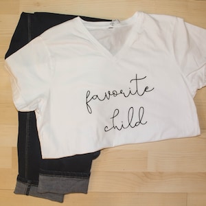 May include: White t-shirt with black text that says "favorite child" on a wooden surface.