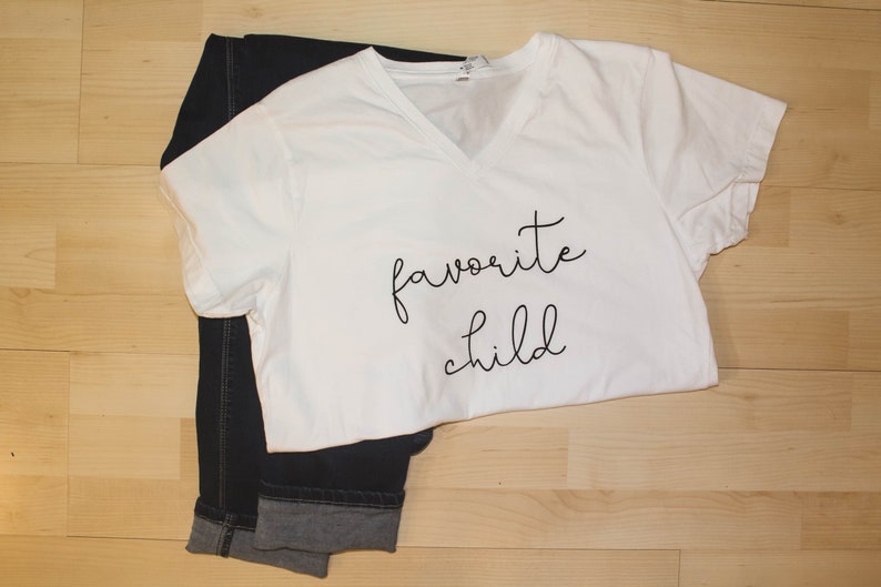 May include: White t-shirt with black text that says "favorite child" on a wooden surface.
