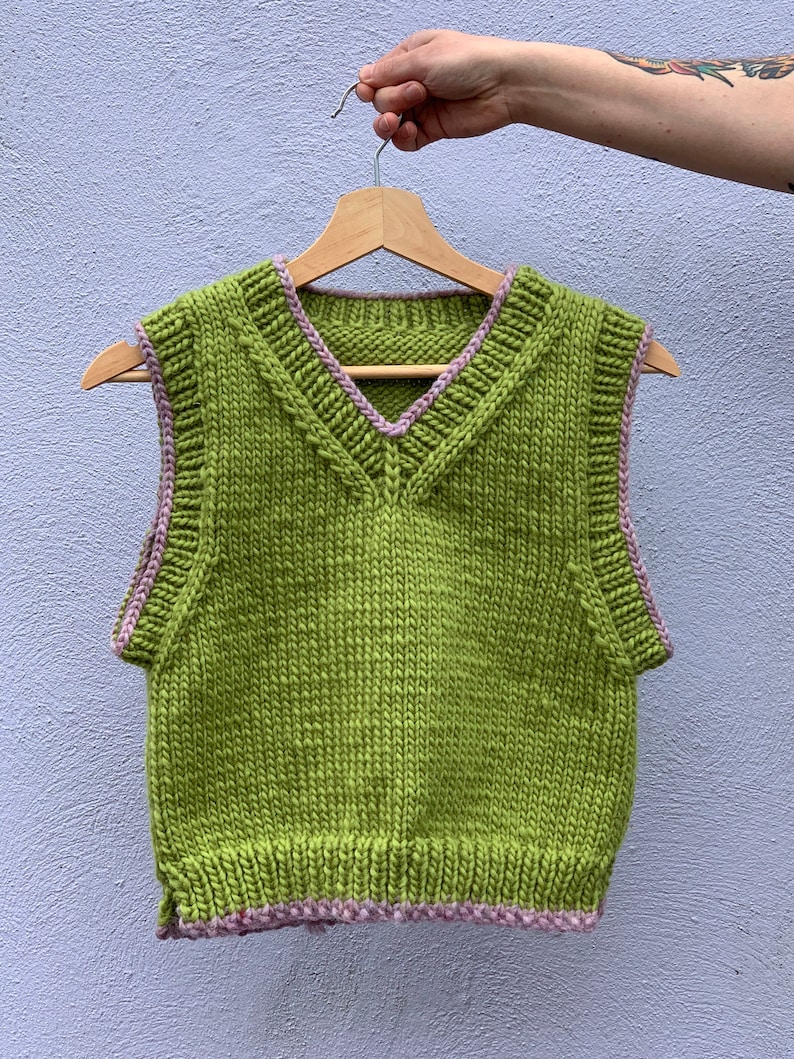 Puede incluir: Un chaleco de punto verde sin mangas con ribete rosa alrededor del cuello y las sisas.