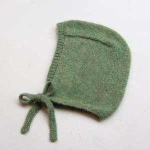 May include: A green knitted baby bonnet with a tie at the back.