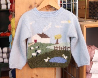 May include: A light blue knitted sweater with a colorful embroidered scene of a house, trees, sheep, and a pond. The sweater is hanging on a wooden hanger.