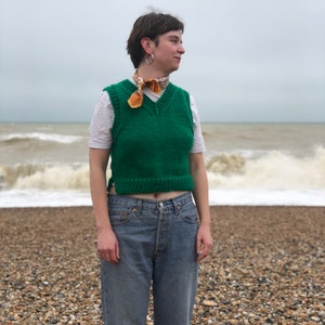 Puede incluir: Una persona lleva un chaleco de punto verde sobre una camiseta blanca, pantalones vaqueros azules y una bufanda estampada. La persona est&aacute; de pie en una playa con olas de fondo.