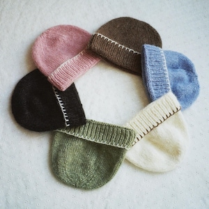 Puede incluir: Seis gorros de punto de diferentes colores, incluyendo marrón, rosa, azul, negro, verde y blanco. Los gorros están dispuestos en círculo sobre un fondo blanco.