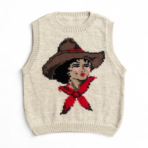 Puede incluir: Un chaleco beige tejido con un diseño de cara de vaquera. La vaquera tiene un sombrero marrón, cabello negro y un pañuelo rojo. El chaleco sin mangas tiene un cuello redondo, perfecto para combinar.