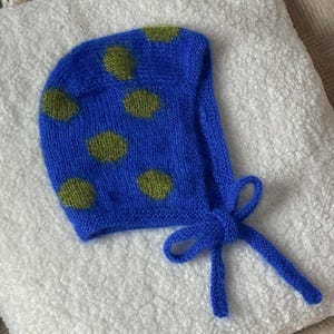 May include: A bright blue knitted bonnet featuring a pattern of fuzzy olive green polka dots. The headwear is constructed with a soft, textured yarn and includes two long blue knitted ties that are knotted together at the front for closure.