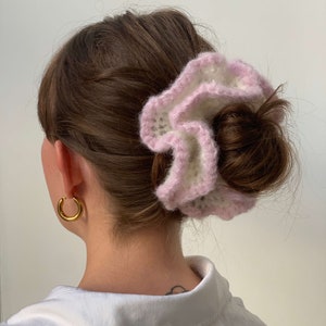 Könnte beinhalten: Ein rosa-weißes gehäkeltes Haargummi mit Rüschen. Das Haargummi wird in den Haaren einer Frau getragen, die zu einem Dutt gebunden sind.