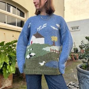 May include: A light blue knit sweater with a landscape design. The sweater features a house, sheep, and a pond. The sleeves are a solid light blue. The sweater is worn with blue and white striped pants.