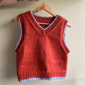 Puede incluir: Un chaleco de punto de color &oacute;xido con cuello en V y ribete azul alrededor del cuello y las sisas.