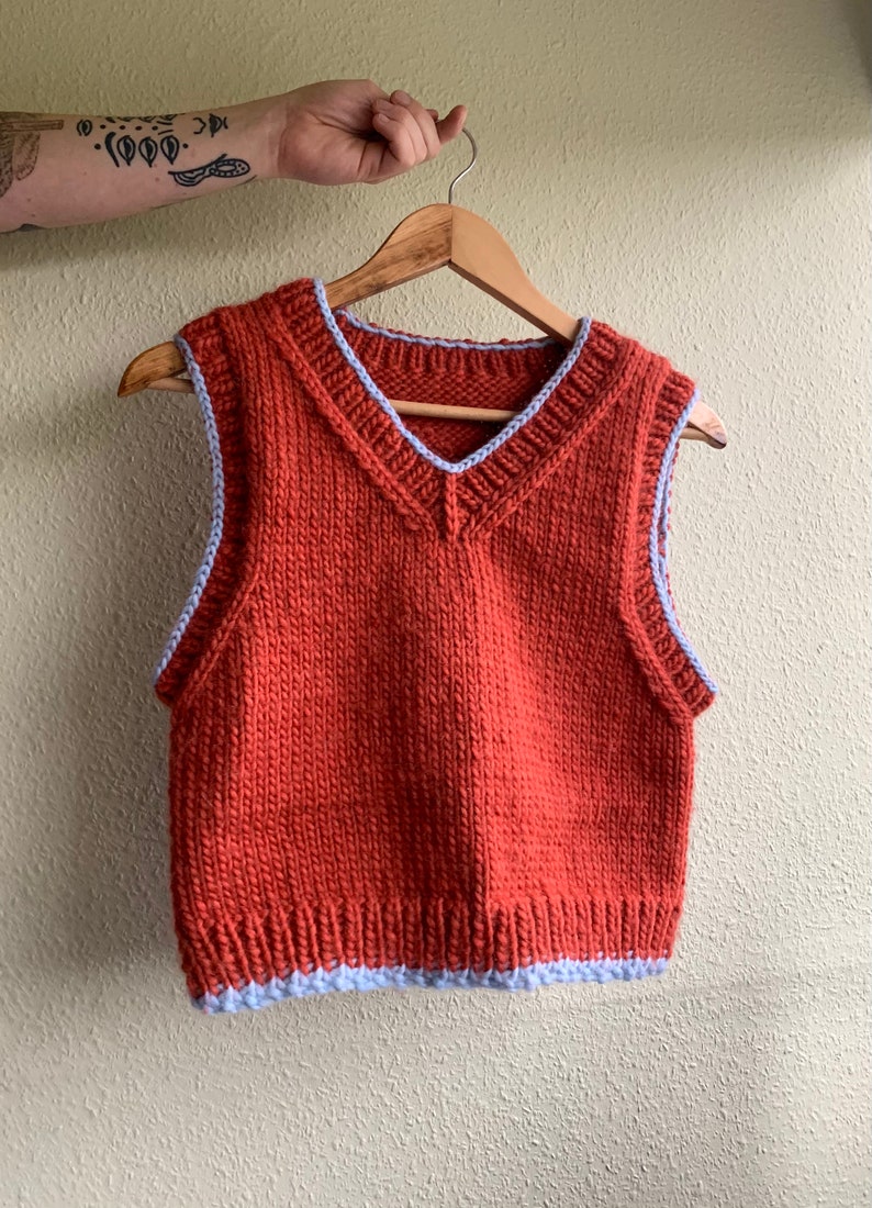 Puede incluir: Un chaleco de punto de color &oacute;xido con cuello en V y ribete azul alrededor del cuello y las sisas.