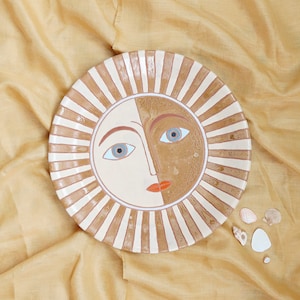 Peut inclure: Une assiette en céramique avec un motif de visage de soleil. Le soleil a un visage blanc avec des accents bruns et orange. L'assiette a une bordure rayée brune et blanche.